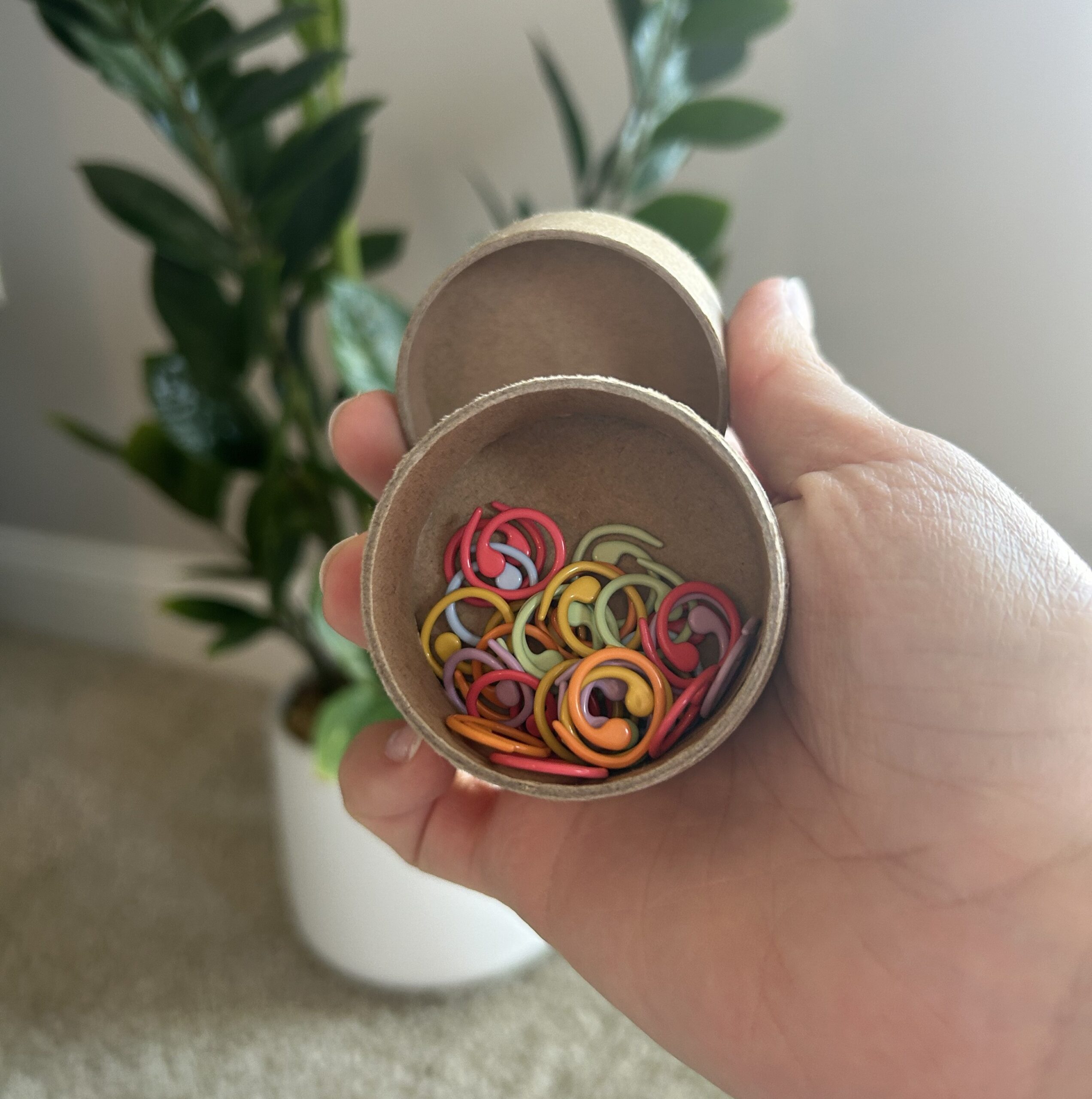 A hand holding an open round container filled with colorful crochet stitch markers in red, orange, yellow, and green