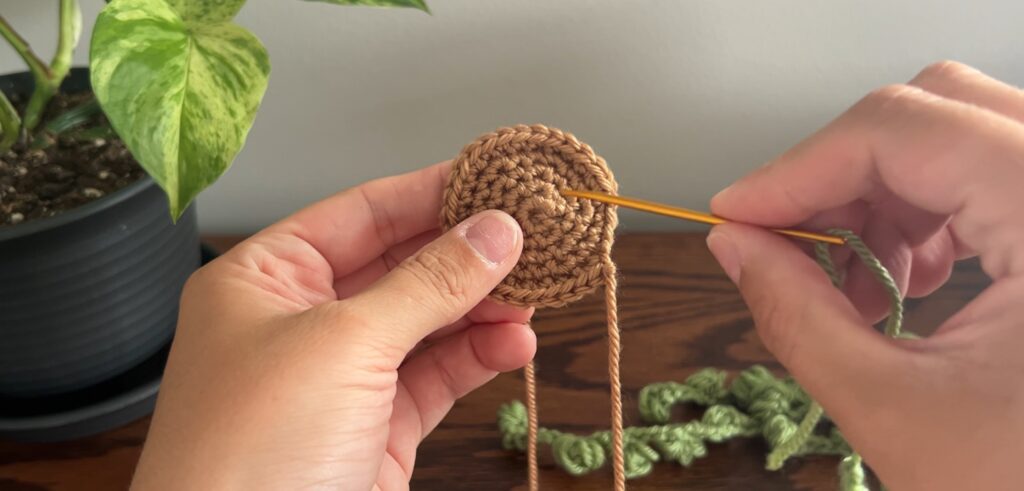 Hand holding a circular crochet circle and darning needle feeding green crochet bulbs through the piece to tie on the opposite side.