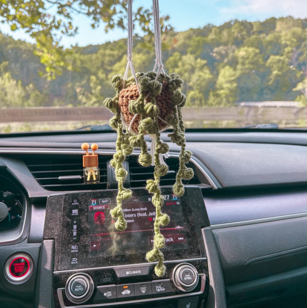 Crochet green plant car accessory hanging from white macrame holder attached to rearview mirror