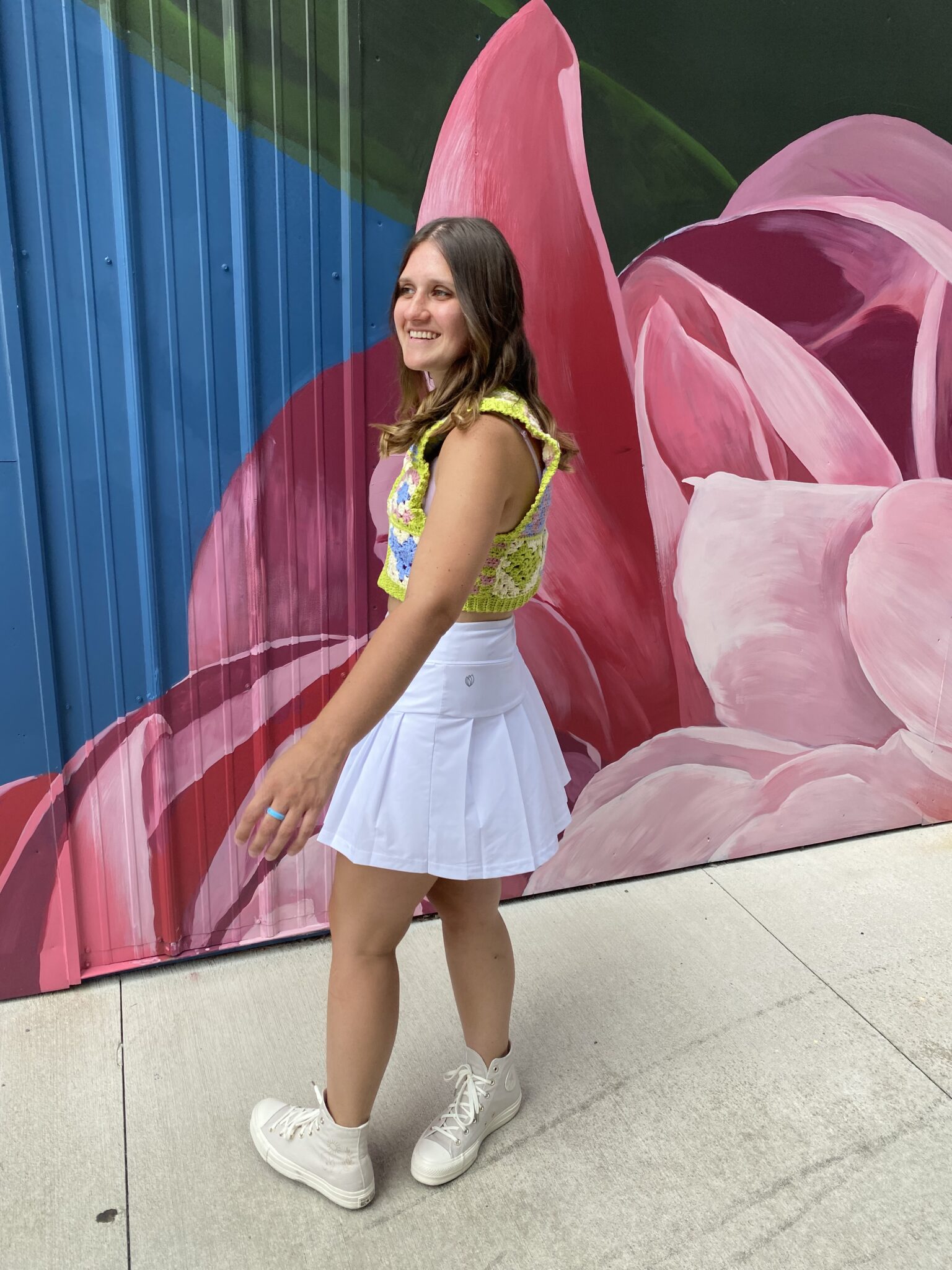 Woman wearing a handmade granny square crochet tank top in green, blue, and pink paired with a white pleated skirt and white sneakers