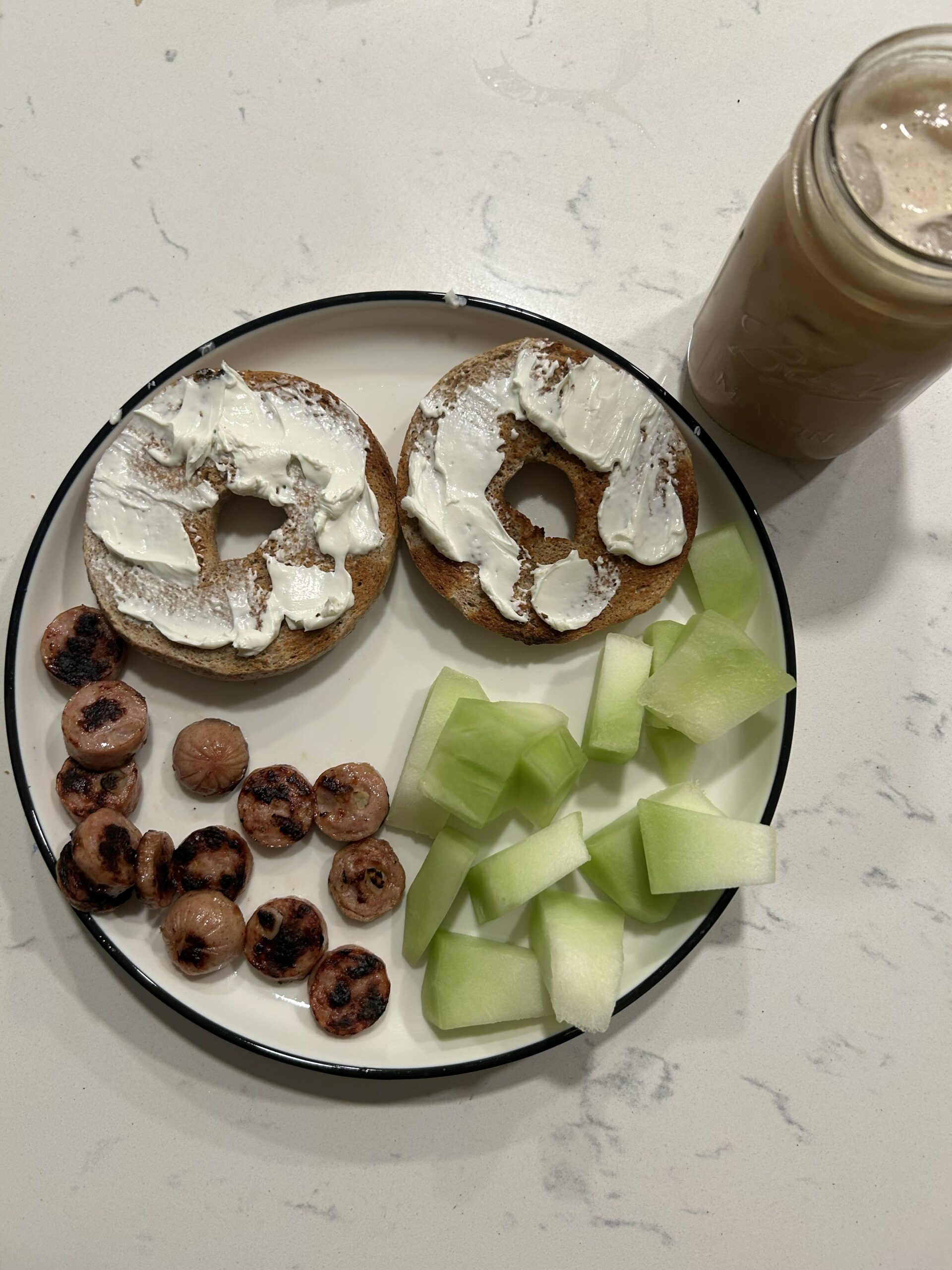 Bagel with cream cheese, circle sausage and honeydew on white plate on marble countertop