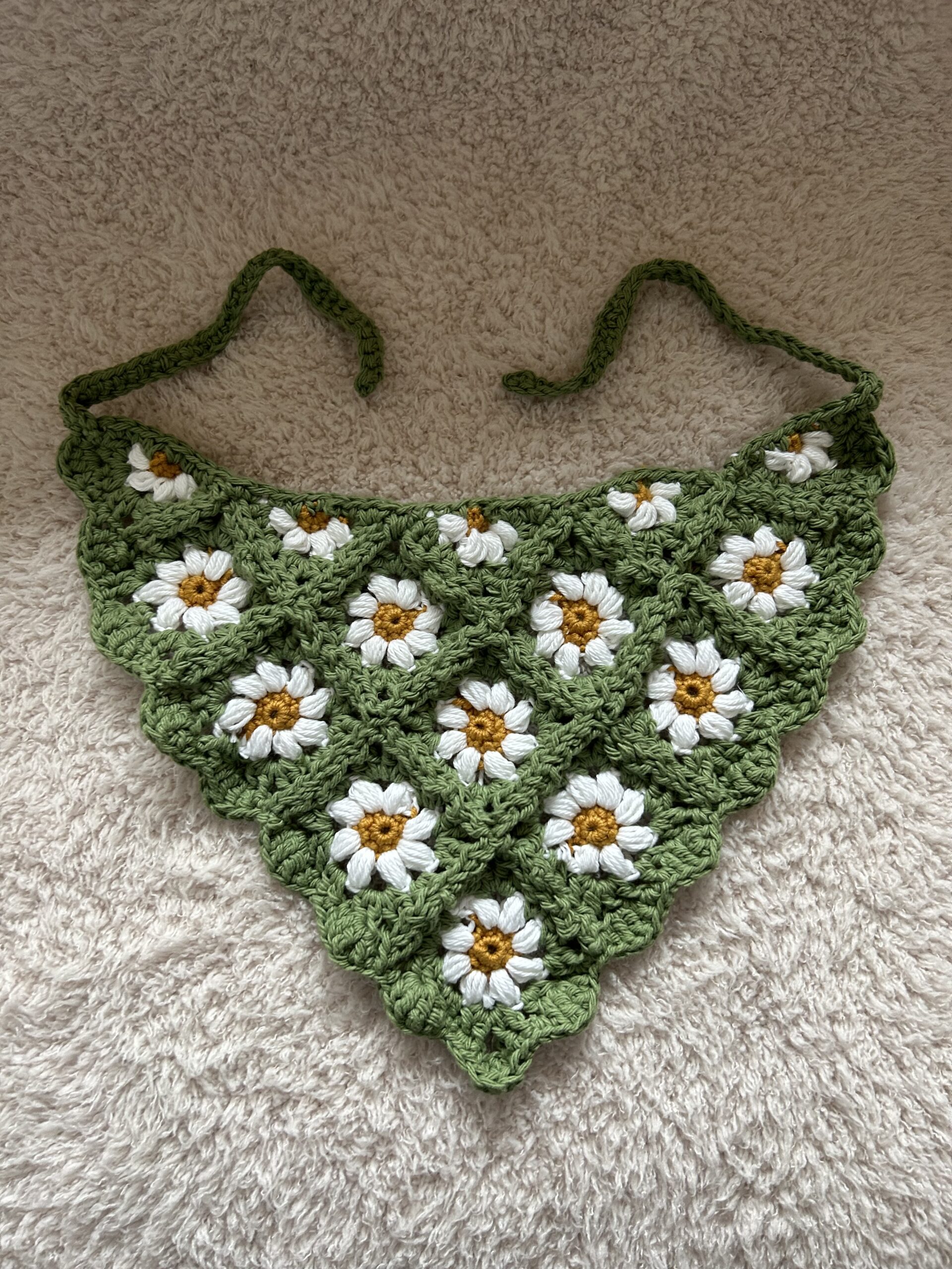 A handmade crochet headscarf with sage green and daisy flowers laying on a light fuzzy blanket