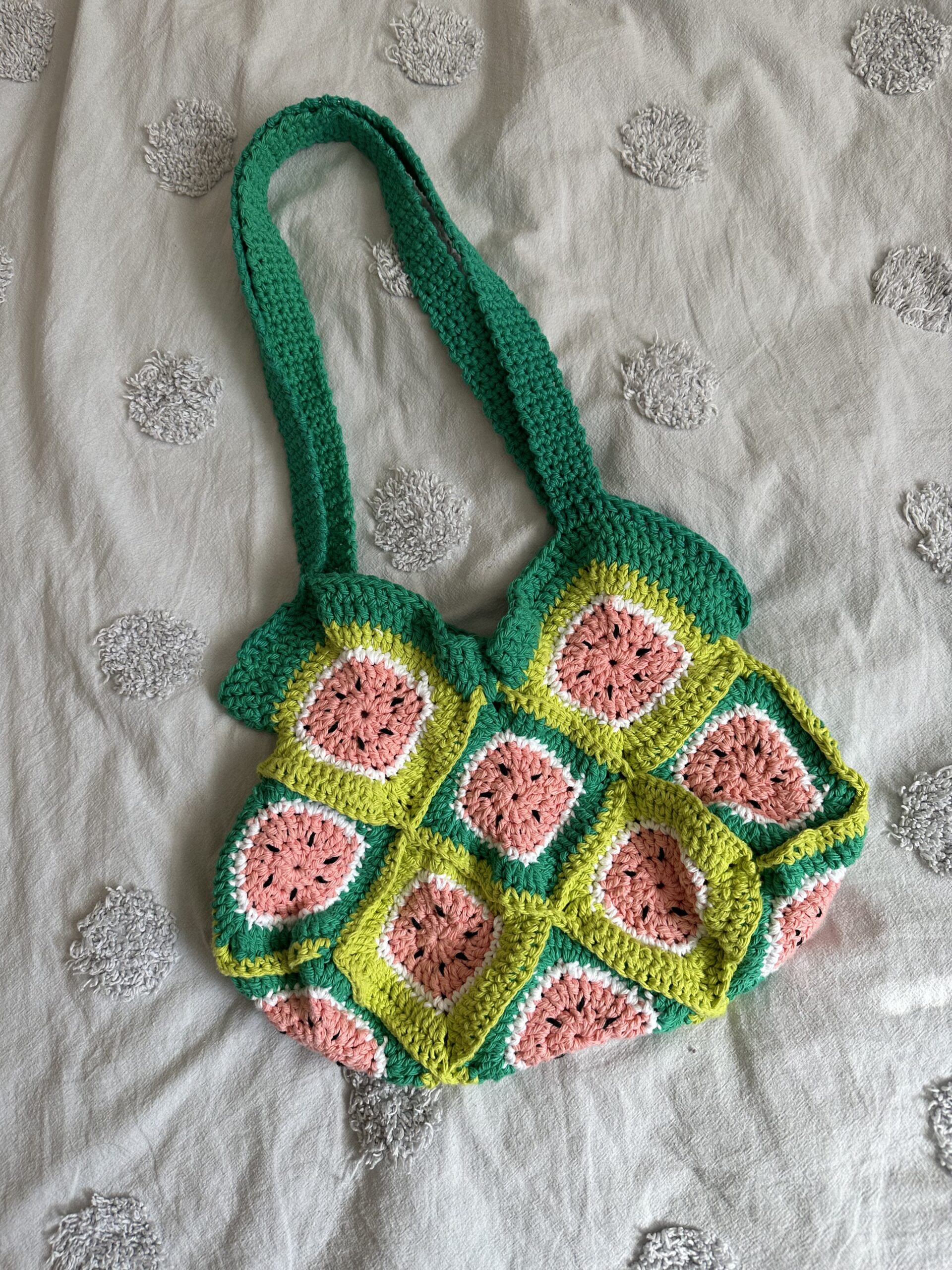 Handmade crochet tote bag with green, pink and black colors to resemble watermelon granny squares laying on light grey bed cover