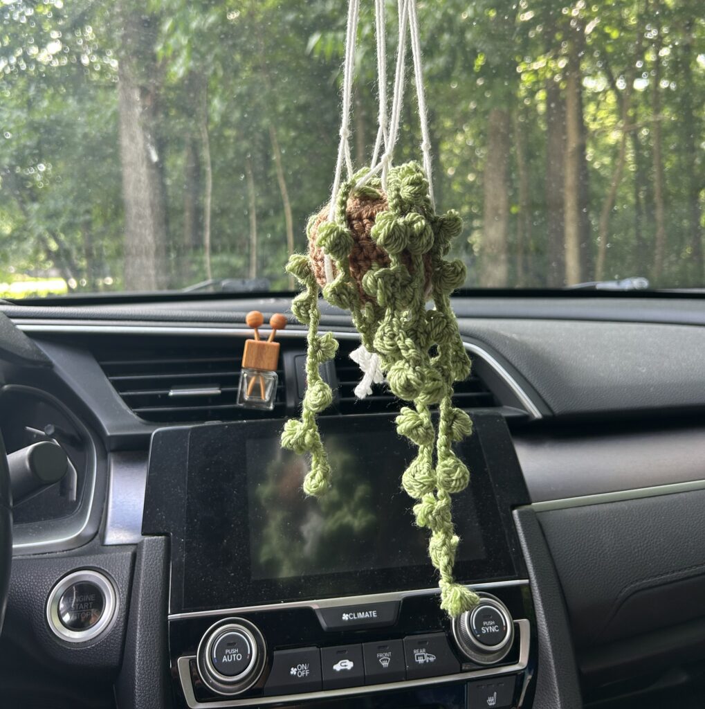 green crochet string of pearls plant sitting on a light brown crochet pot in a white macrame holder hanging from the rearview mirror in a sedan car