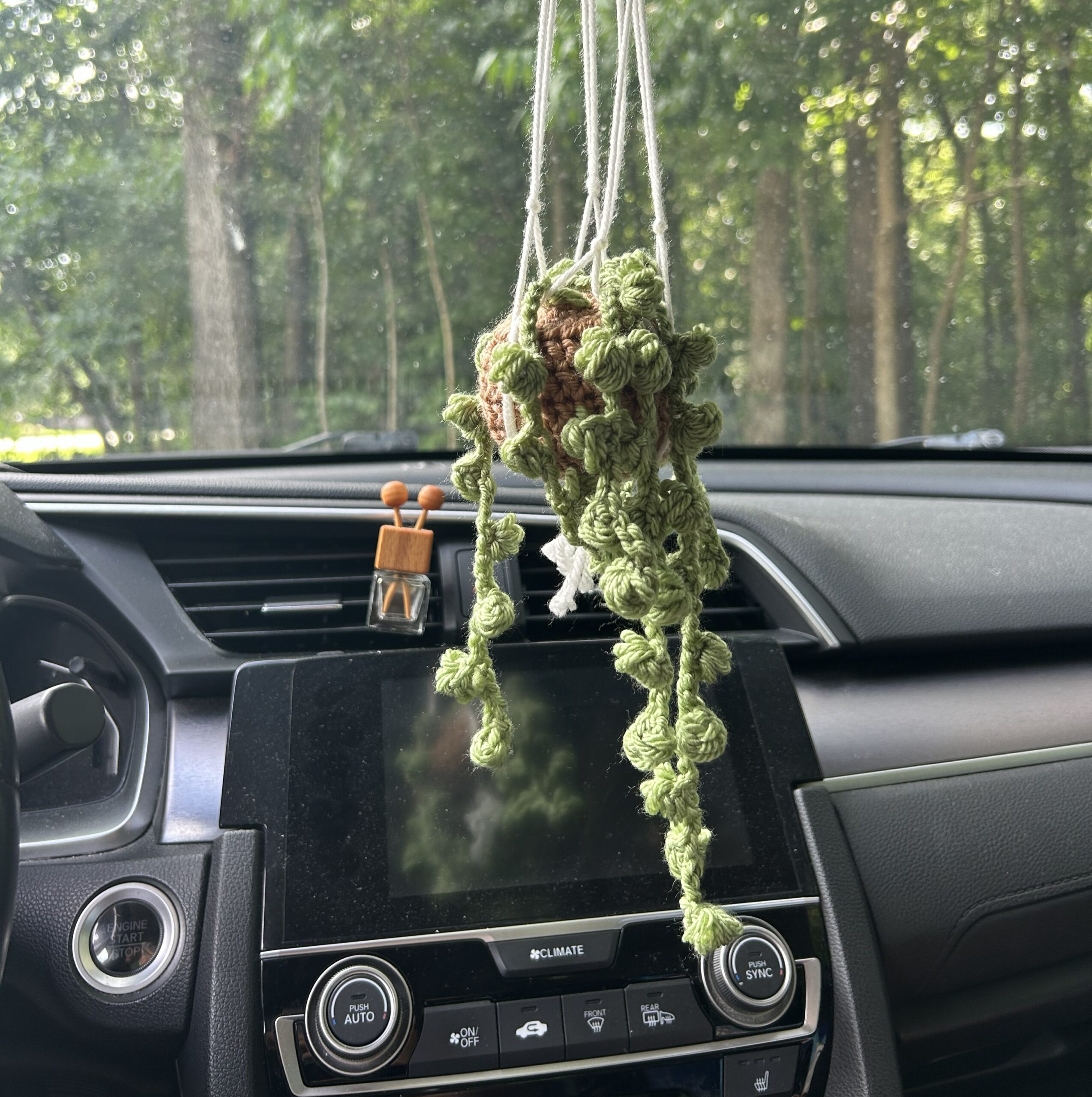 green crochet string of pearls plant sitting on a light brown crochet pot in a white macrame holder hanging from the rearview mirror in a sedan car
