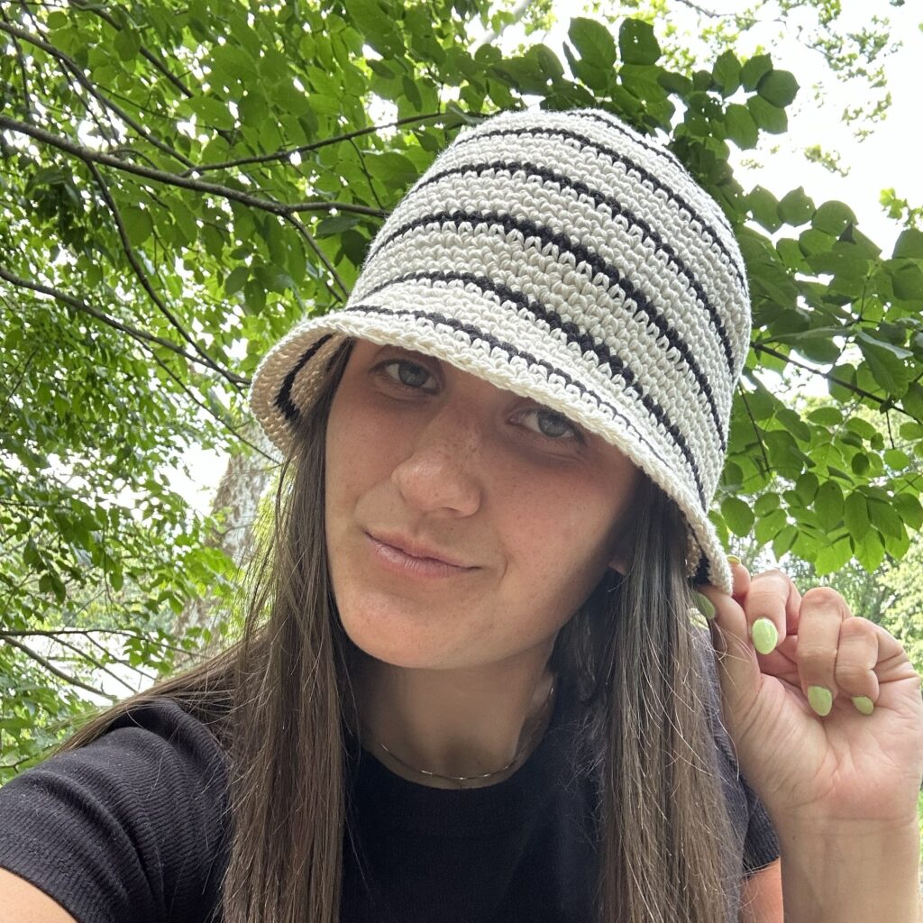 Young woman with brunette hair taking a selfie while wearing a black and white crochet bucket hat with black shirt on.