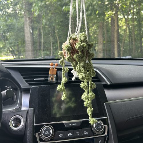 green crochet string of pearls plant sitting on a light brown crochet pot in a white macrame holder hanging from the rearview mirror in a sedan car